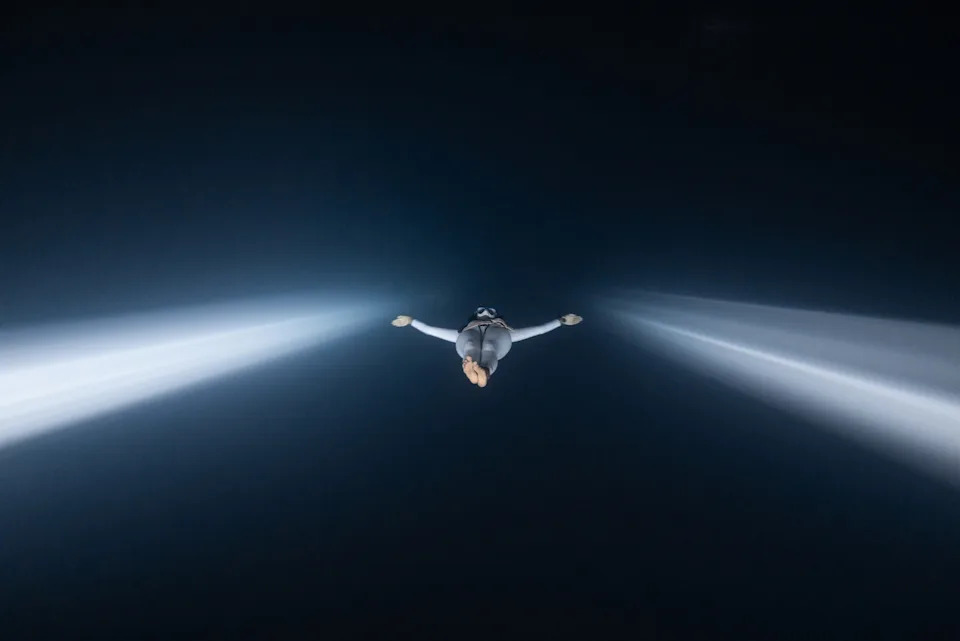 A freediver, seen deep underwater, flanked by two shafts of light extending into the dark water beyond.