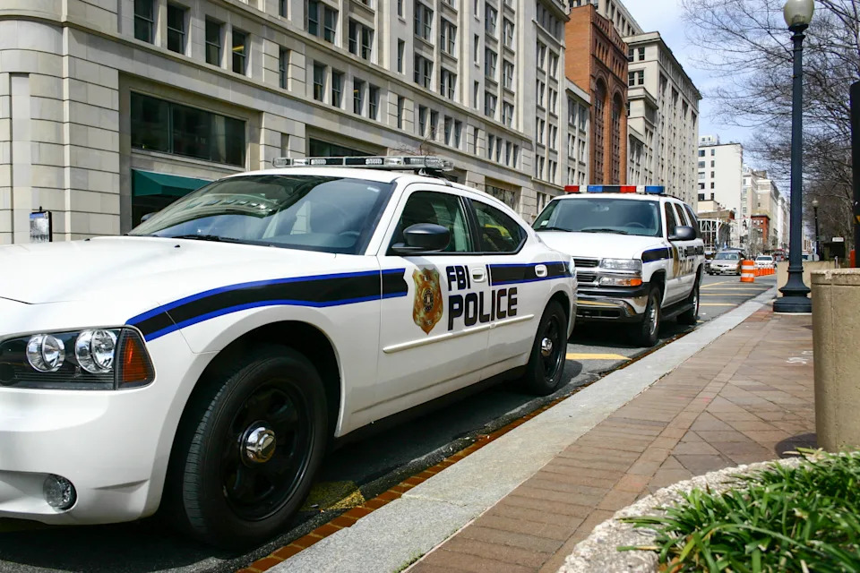 FBI police car parked on a city street, with another police vehicle in the background, near a line of buildings