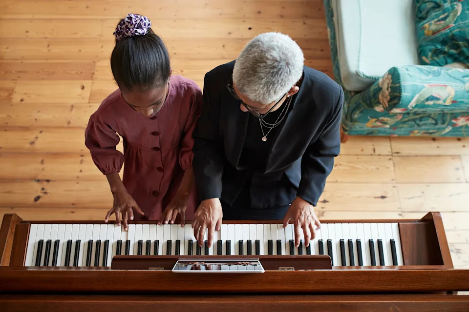 A child in a dress and adult in a blazer play piano together, viewed from above, highlighting a shared musical moment