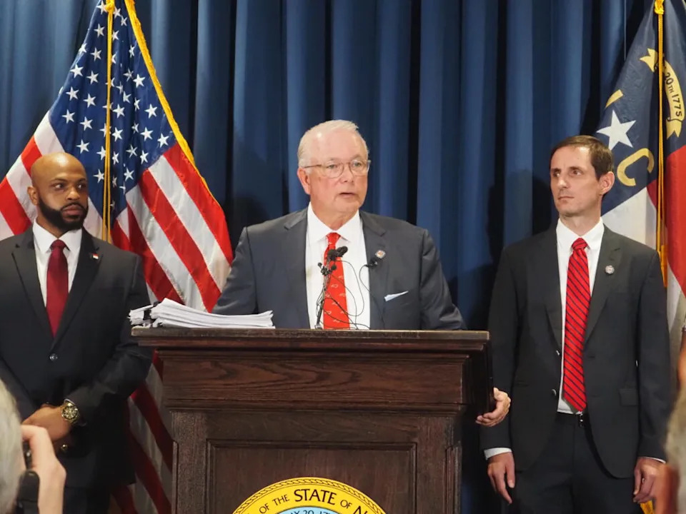 Dave Boliek speaks at a podium next to the flags of the United States and North Carolina.
