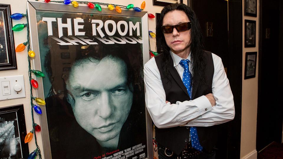 Tommy Wiseau, con cabello largo y negro, gafas negras y un chaleco negro, se apoya en un póster de