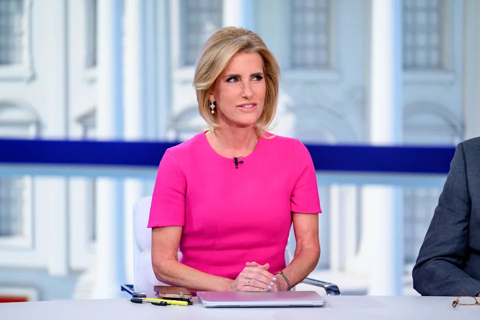 Person seated at a news desk, wearing a short-sleeved dress, engaged in discussion