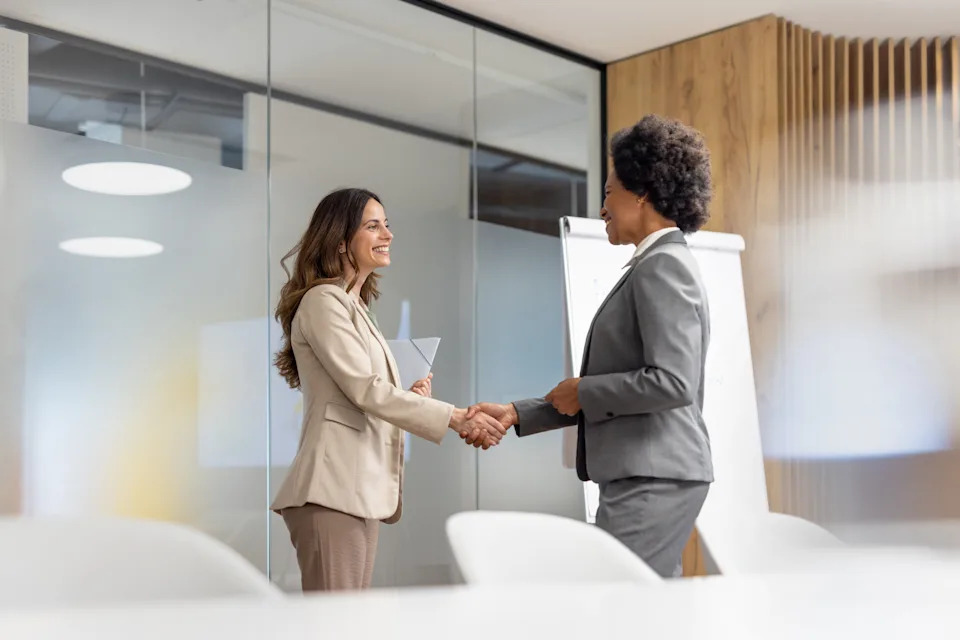 Two professionals in business attire are smiling and shaking hands in a modern office setting