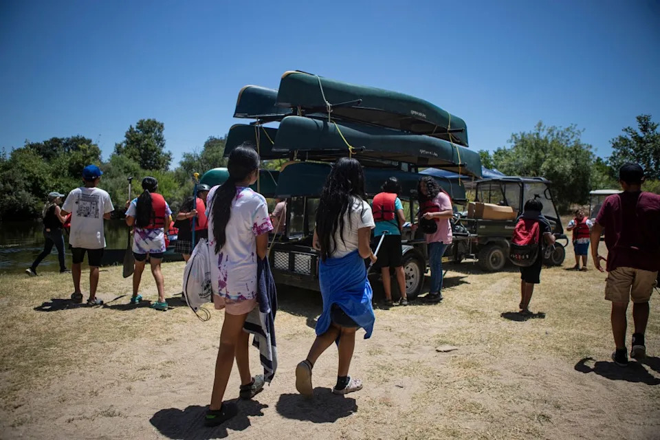 A group of children walking around a stack of upside-down canoes piled on a rack that is hitched to the back of a golf cart and parked next to a riverbed.