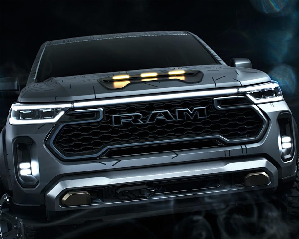 Front view of a gray RAM truck with illuminated LED headlights and a prominent black grille featuring the RAM logo. The hood has three amber marker lights, and the bumper includes metallic accents and fog lights. The background is dark with subtle smoke effects.