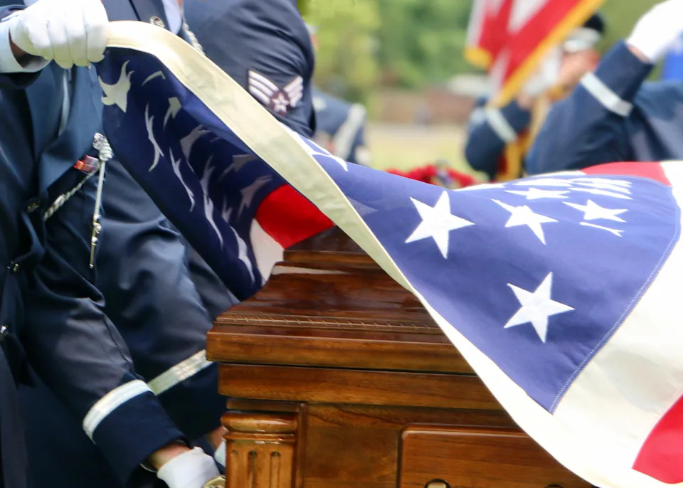Military personnel drape an American flag over a wooden casket during a ceremonial event
