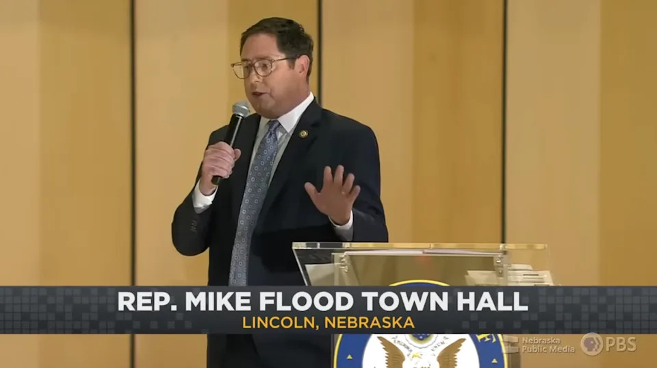 A man is speaking at a podium with "Rep. Mike Flood Town Hall, Lincoln, Nebraska" displayed on the screen