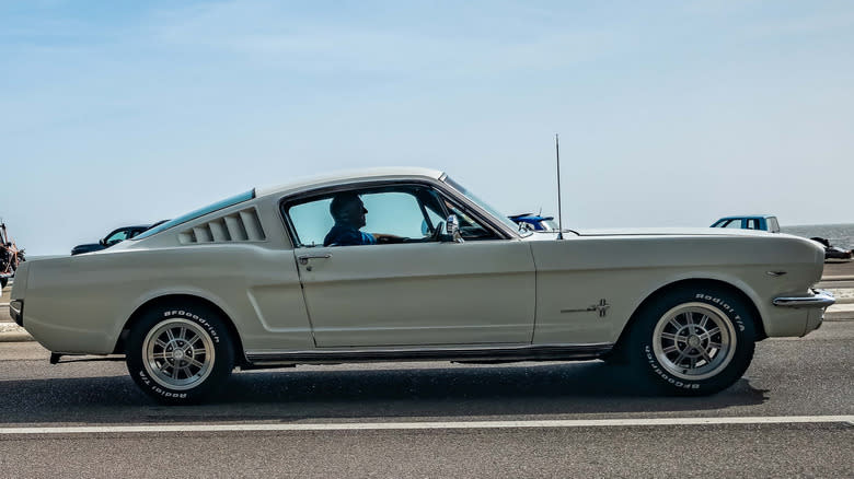 A white 1965 Ford Mustang with a fastback body style driving on the road.