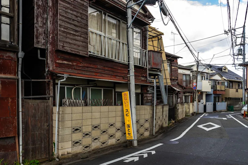 Narrow street in a residential area with older wooden houses and a yellow safety sign in Japanese near a street corner