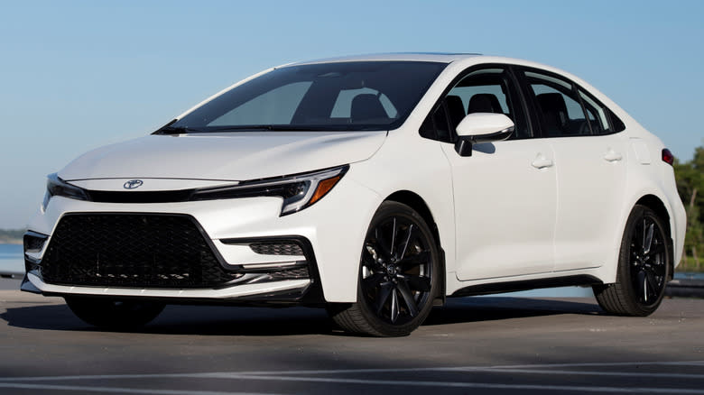 A Wind Chill Pearl 2023 Toyota Corolla XSE parked on a paved surface with trees and a body of water in the background