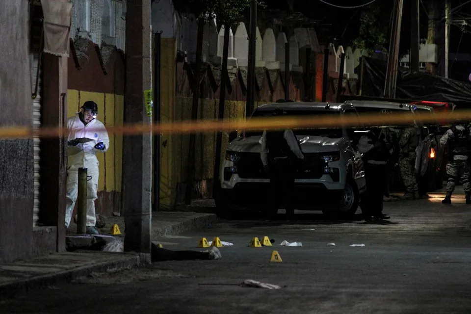 Forensic personnel work behind crime-scene tape in a street.