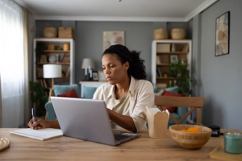 A person works on a laptop at a dining table with an open notebook, takeout container, and bowl of oranges nearby, in a cozy living room