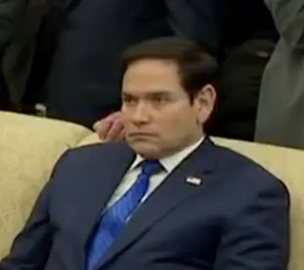 Person in a suit seated on a couch, looking serious, in a formal setting with people gathered around