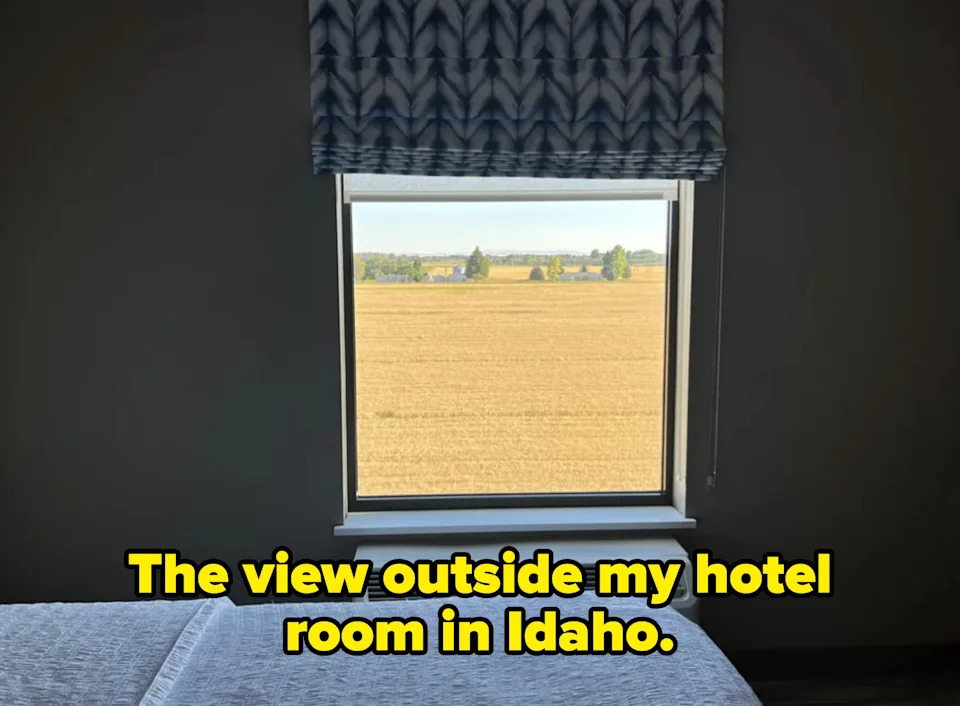 A window view of a flat, expansive field under a clear sky, seen from inside a room with a bed. Caption: "The view outside my hotel room in Idaho"