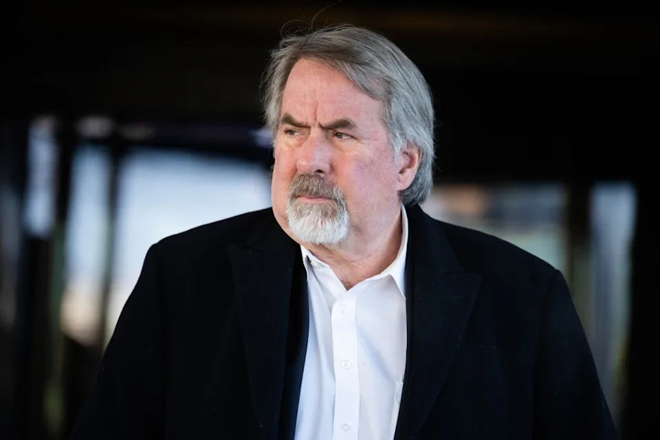 A person with a beard and mustache is wearing a white shirt and dark jacket, looking to the side with a serious expression