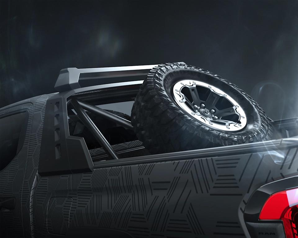 Close-up of the rear section of a black pickup truck with a geometric pattern on the body, featuring a mounted spare off-road tire with a rugged tread and a silver and black alloy wheel. The truck bed has a black roll bar, and part of the red tail light is visible. The background is dark and smoky.