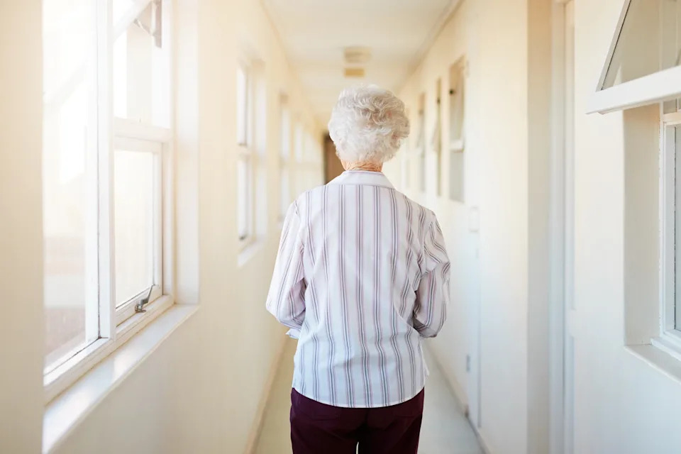 Older person with short hair walks alone down a bright hallway, wearing a striped shirt and dark pants