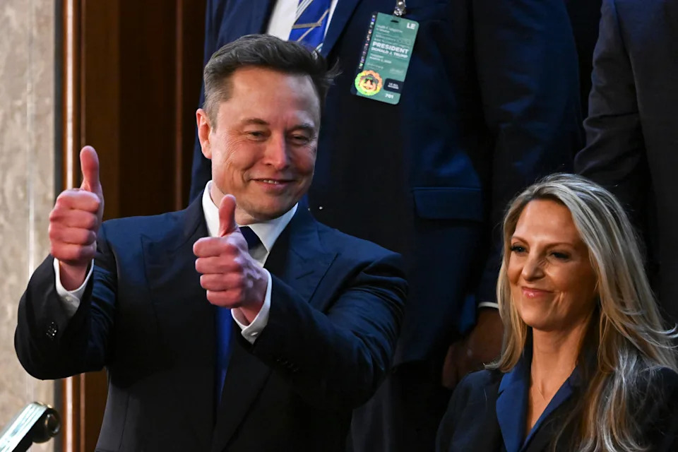 A person in a suit gives two thumbs up while standing next to another person smiling