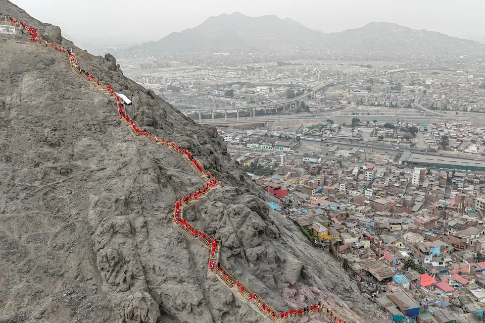 A long line of people attending a memorial are seen standing on a winding path on the side of a rocky mountain.