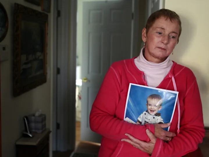 A woman in a casual outfit holds a framed photo of a smiling baby, standing thoughtfully in a domestic setting