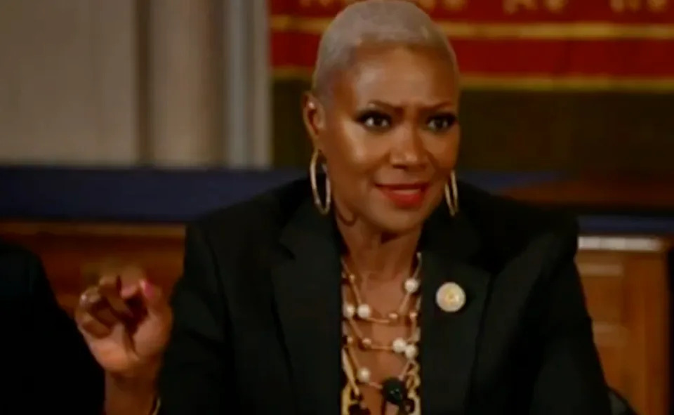 A person in a black blazer and pearl necklace gestures while speaking at a formal event