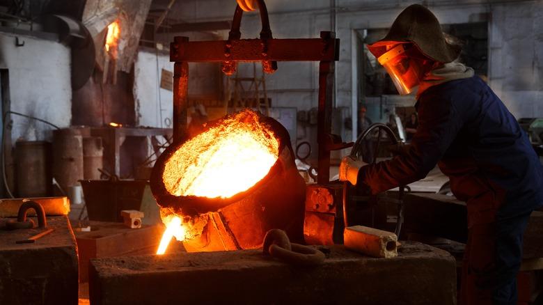 Industrial worker in protective gear pours molten metal at steel foundry. Manufacturing process in heavy industry with furnace. Skilled laborer handles high-temperature metallurgy, safety action.