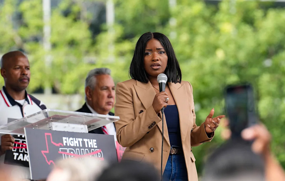 Person speaking at an outdoor event, holding a microphone. Others stand nearby, with a "Fight the" sign partially visible in the background