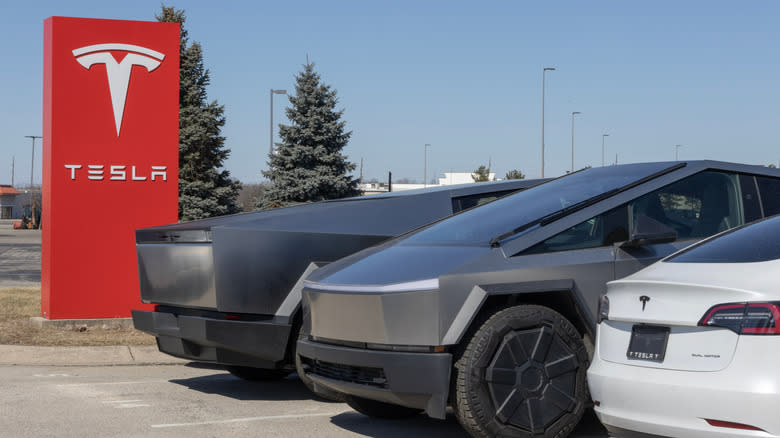Three tesla cars, two cybertrucks and a Model 3, parked in front of a red Tesla sign