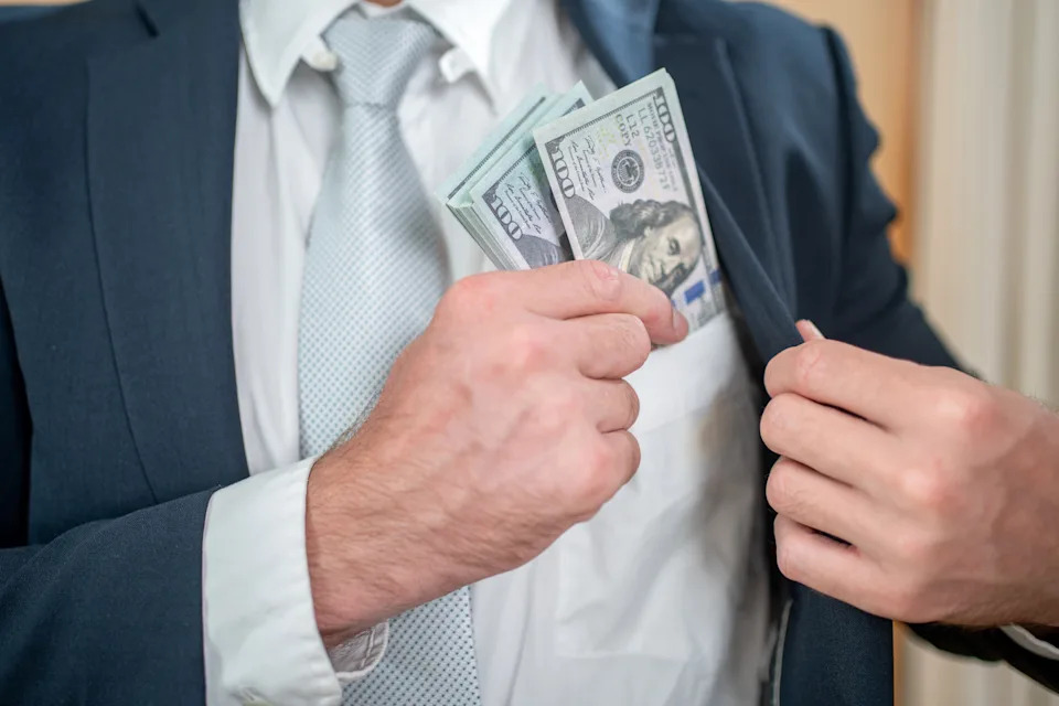 Person in a suit placing a stack of hundred-dollar bills into their jacket pocket. Only torso and hands are visible