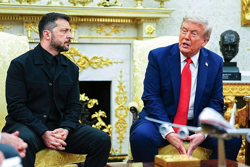Two men sitting in an ornate room engaged in conversation, one in a black suit and the other in a blue suit with a red tie