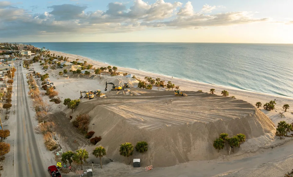 Aerial view of beach area with large sand pile, construction machinery, and a road lined with palm trees running parallel to the coastline