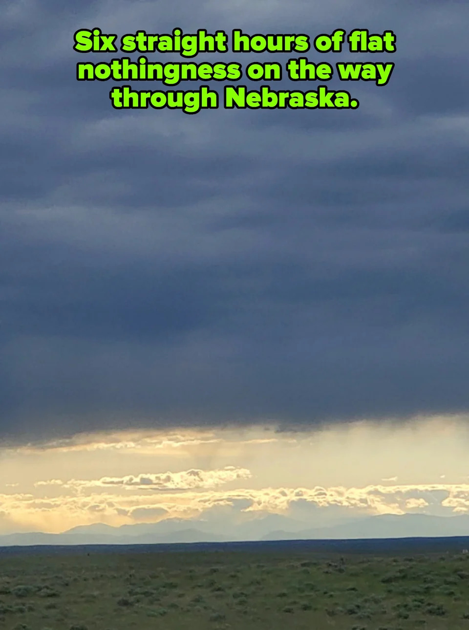 Cloudy sky over distant mountains with a hint of sunlight peeking through, casting light on a vast landscape. Caption: "Six straight hours of flat nothingness on the way through Nebraska"