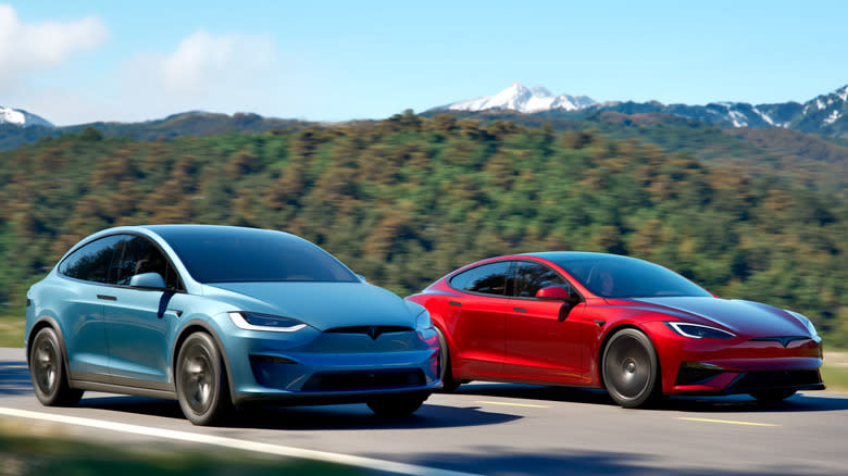 a blue Model X driving next to a red Model S in front of trees and mountians