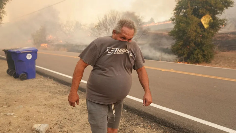 A man walks with a shirt over his nose and mouth while a wildfire burns behind him with smoke filling the air