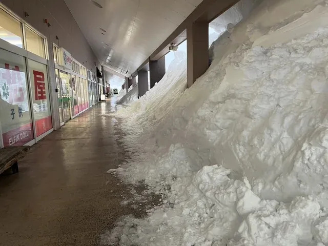 Outdoor shopping area with heavy snow piled against storefronts, partially blocking the walkway