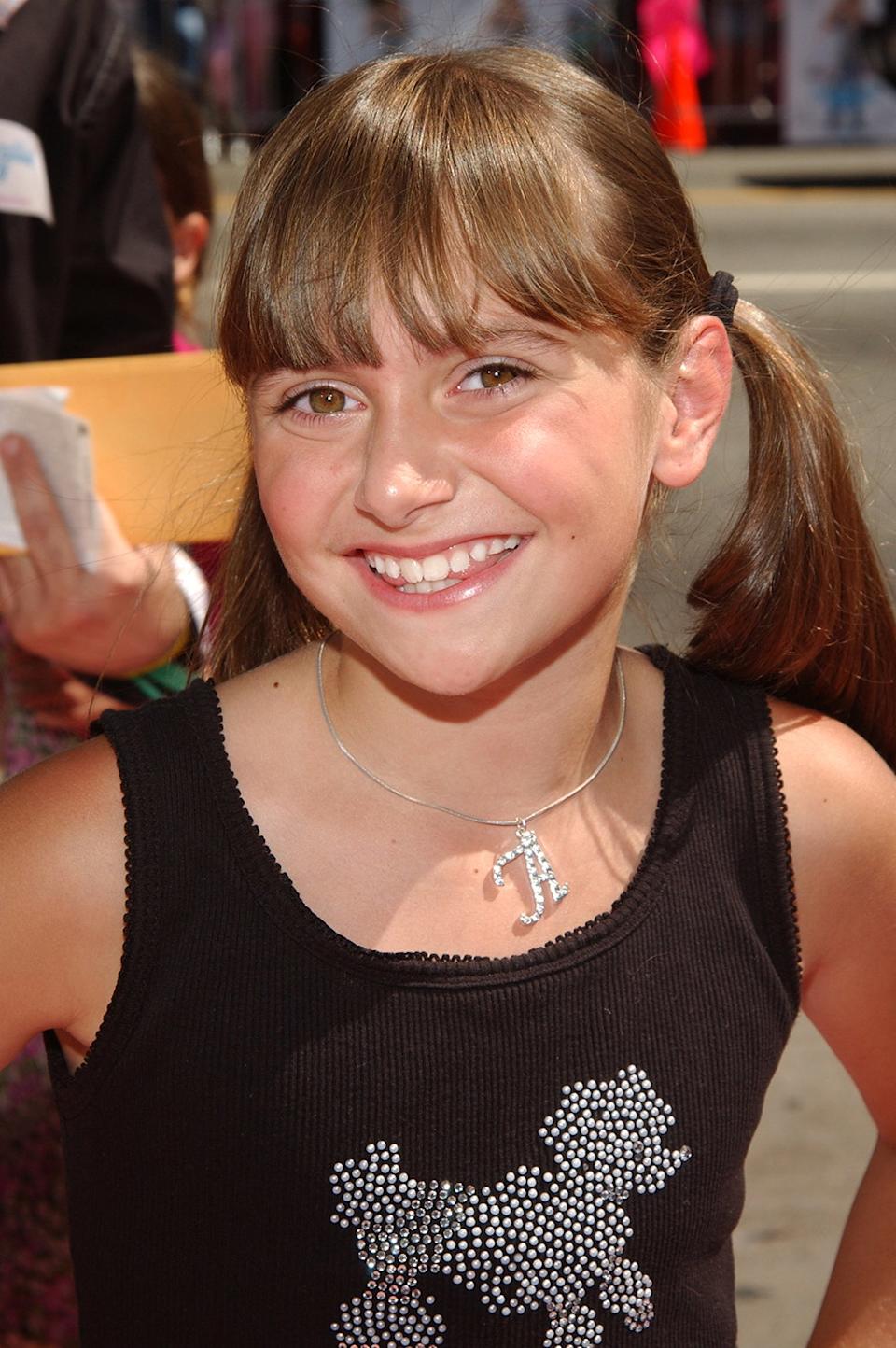 Un primer plano de Alyson Stoner con una camiseta de tirantes negra con un caniche brillante y coletas.