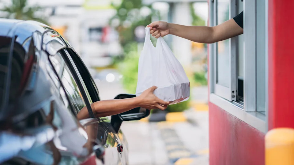 Person in a car receives a takeout bag from a drive-thru window, suggesting a transaction or work scenario