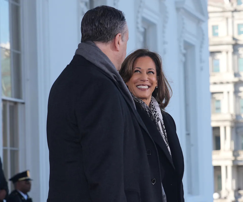 Two people stand outdoors, smiling at each other. One is wearing a dark coat and patterned scarf; the other is in a dark coat with a scarf