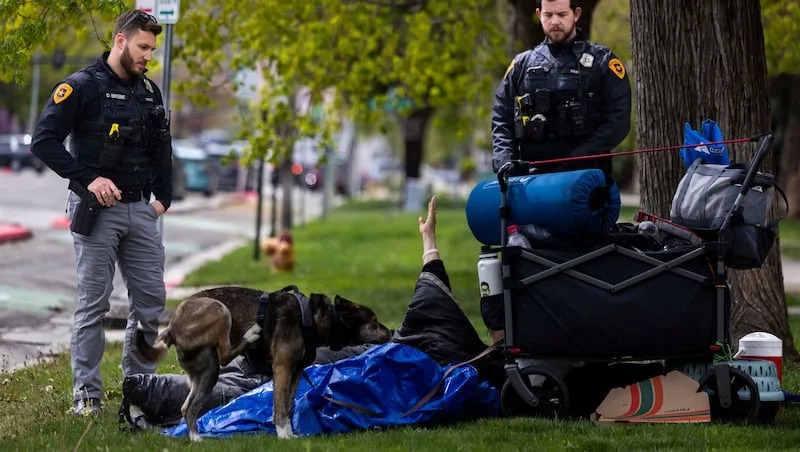 Salt Lake City police Detective Dalton Beebe, left, and Salt Lake City police officer Brad Donahoo, right, inform a homeless man that he can’t sleep on the grass along the sidewalk, in Salt Lake City on Thursday, April 17, 2025.