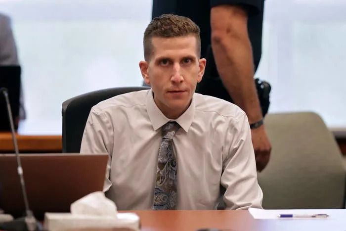 A person in a courtroom sits at a table with a laptop, wearing a patterned tie and light shirt, looking towards the camera