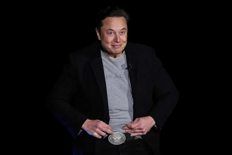 Musk, wearing a black blazer and a gray SpaceX T-shirt, lifts the hem of his shirt to show a large silver belt buckle featuring a five-pointed star with Tesla’s stylized “T” logo in the middle.