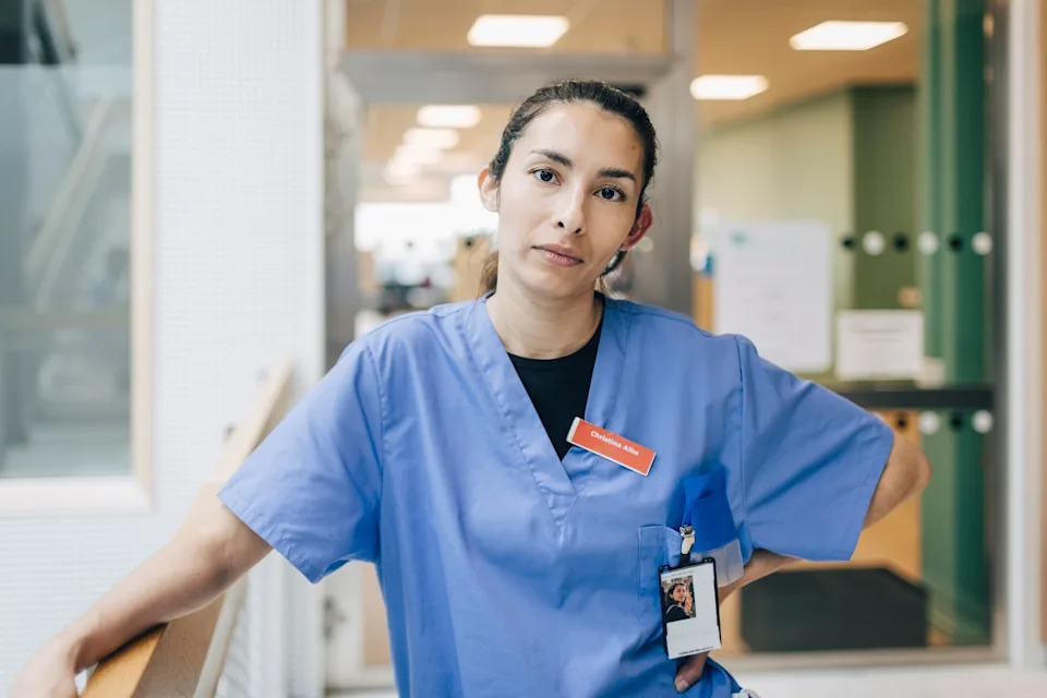 Person in hospital scrubs stands confidently in a medical facility hallway, wearing a staff badge