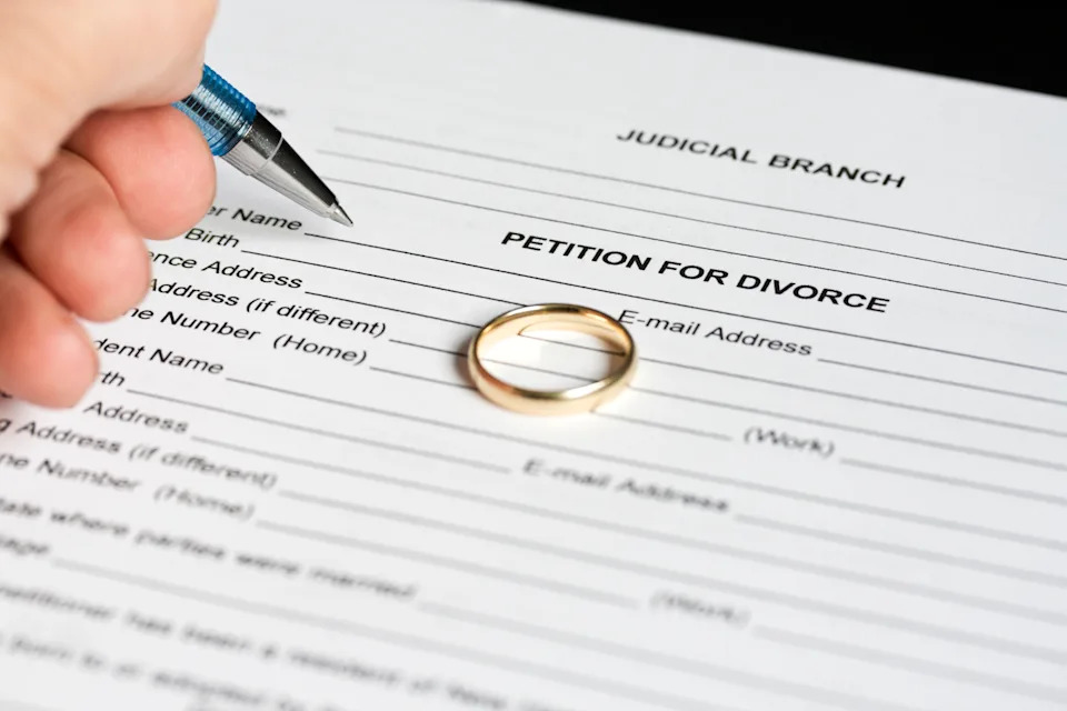 A hand holding a pen is poised over a "Petition for Divorce" form, with a wedding ring placed on the paper
