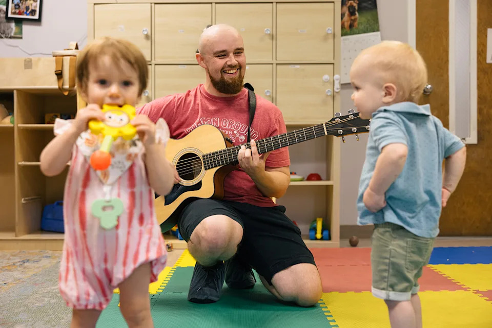 A young man plays guitar for two toddlers in a childcare setting