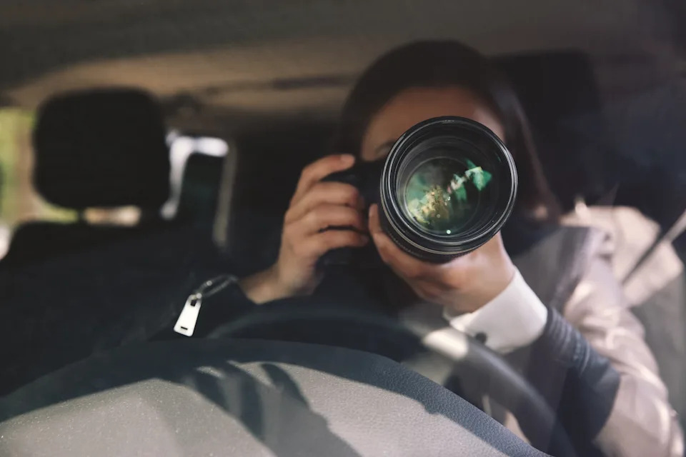 Person holding a camera with a large lens, positioned inside a car, taking a photograph through the windshield