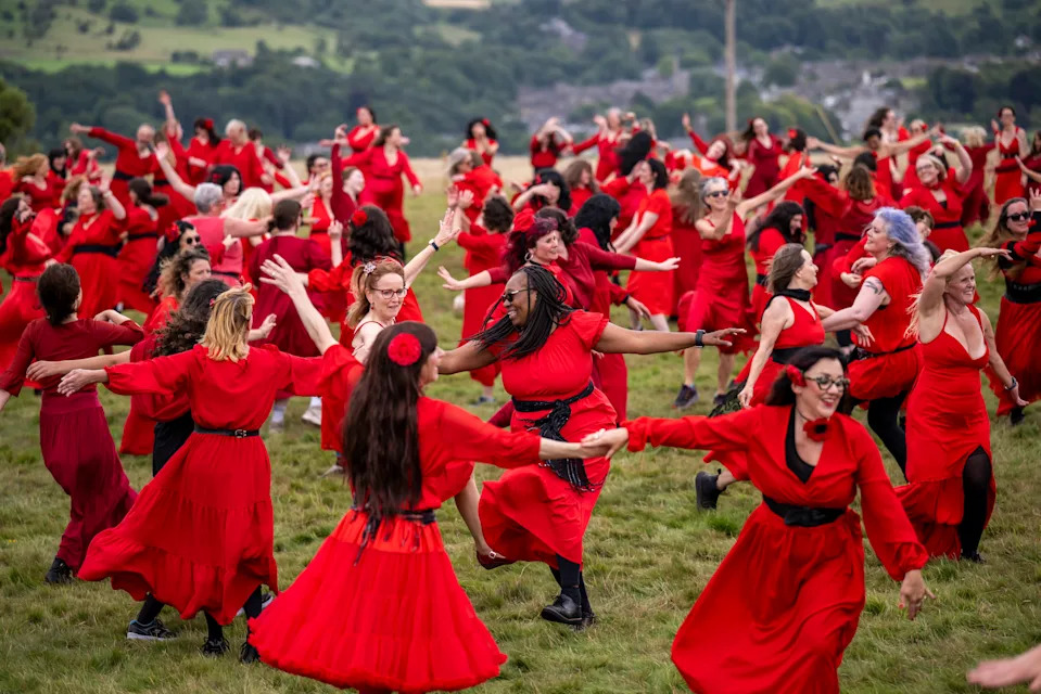 A large group of people, all wearing red dresses, dances in a field.