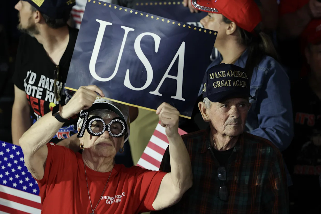 A person in a red t-shirt and a pair of decorative, rhinestone glasses holds up a blue sign that says “USA” in white text. A man looks on in a black “Make America Great Again” hat. They are part of a larger crowd. Behind them is an American flag.