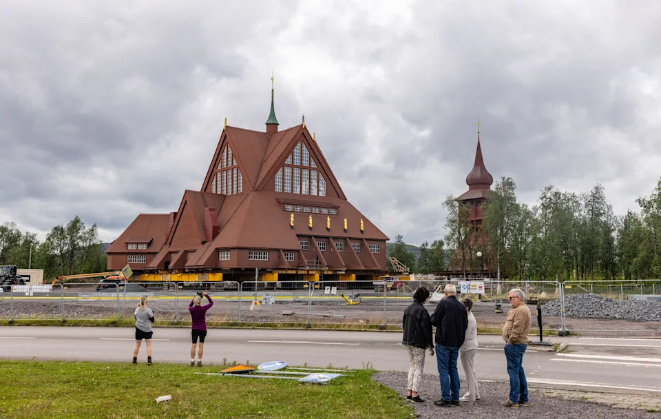 Several people stand near a road, looking at a tall wooden church that has been lifted onto steel beams before a move.