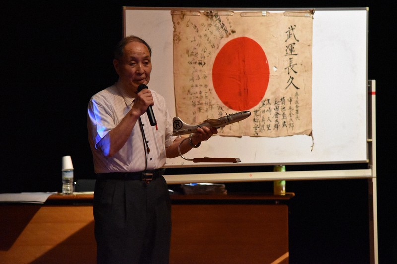 パネルに日章旗を掲げながら講演する小倉さん=栃木市旭町の栃木文化会館で2025年7月10日、太田穣撮影