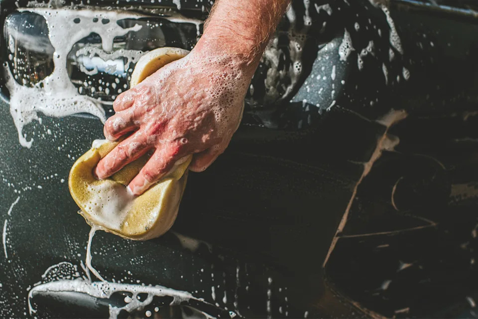 Close-up of a person's hand washing a car with a soapy sponge, highlighting manual labor and effort in car maintenance